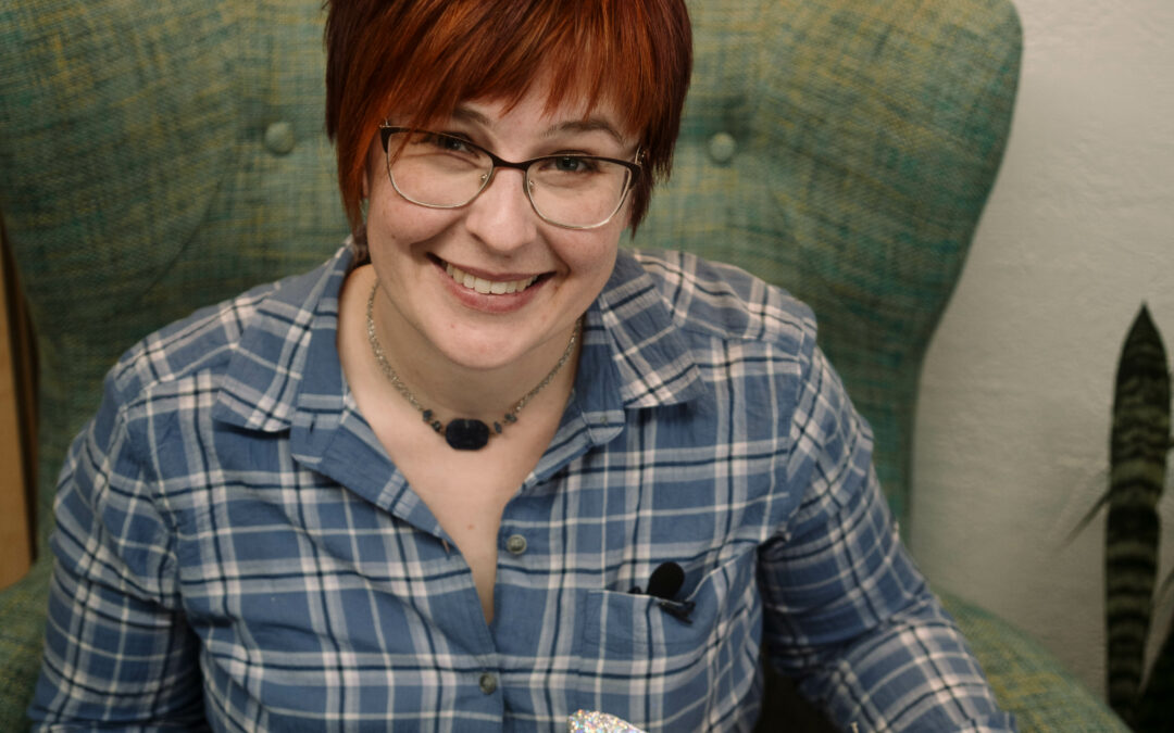 person with short red hair and glasses smiling and holding a pile of messy tissue paper in their lap.