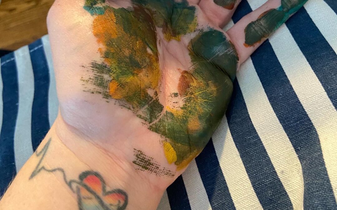 palm of adult with paint on it, tattoo showing a pride heart and sign wave pulse. striped background