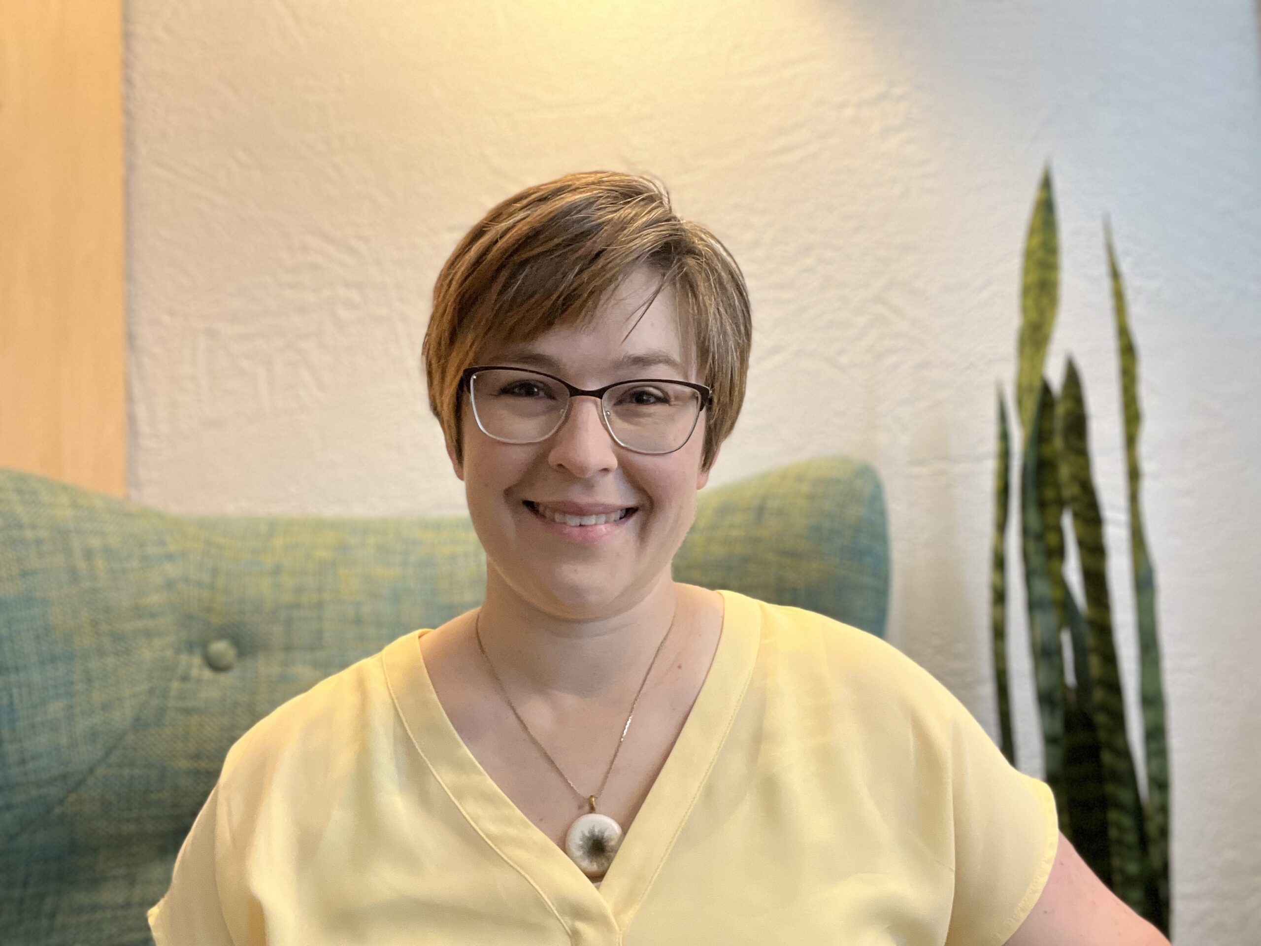 smiling woman with short brown hair and glasses, wearing a  yellow blouse and sitting in a green chair with a snake plant in background