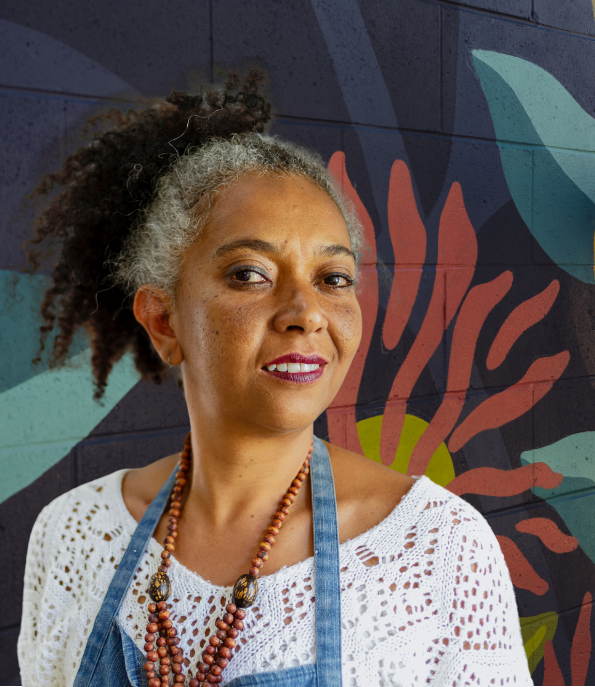 middle aged black woman wearing an artists apron looking confident and happy in front of a mural